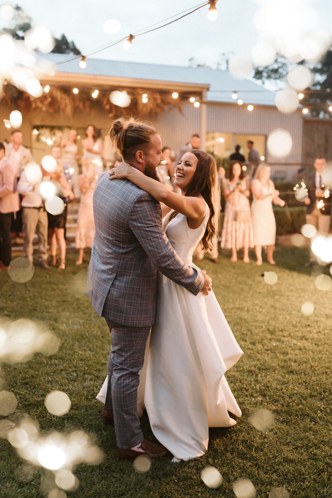 A couple dancing on the lawn under fairy lights, guests behind them