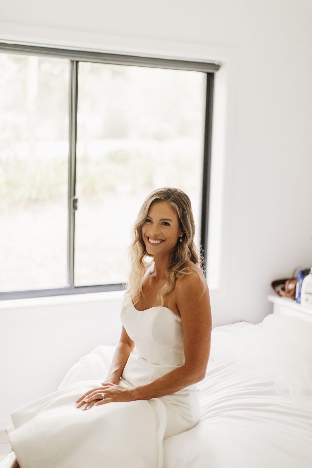 A bride smiling on the bed in a bright cottage bedroom